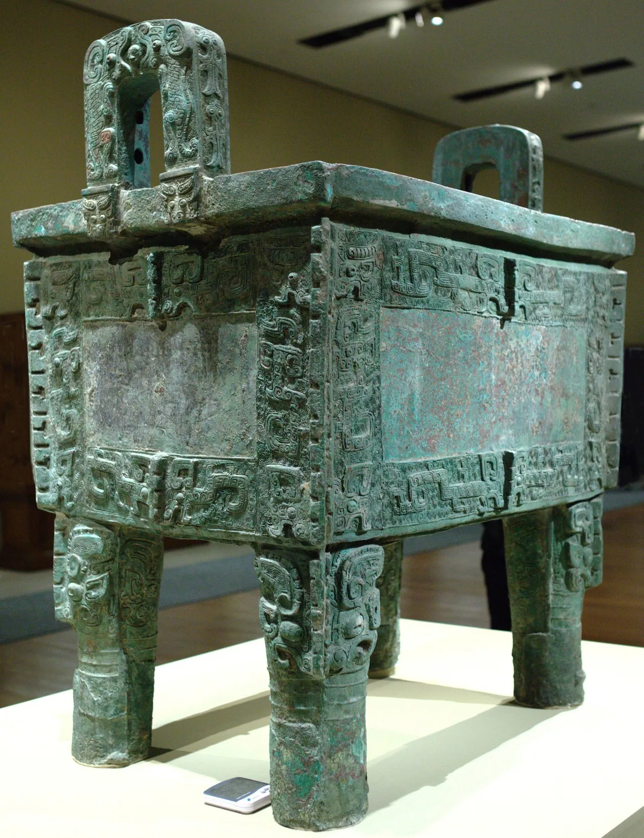 商代后母戊青铜方鼎：四足方鼎立于展柜中，器身布满饕餮纹与盘龙纹，口沿上一对立耳，体量巨大，器表呈深绿色铜锈